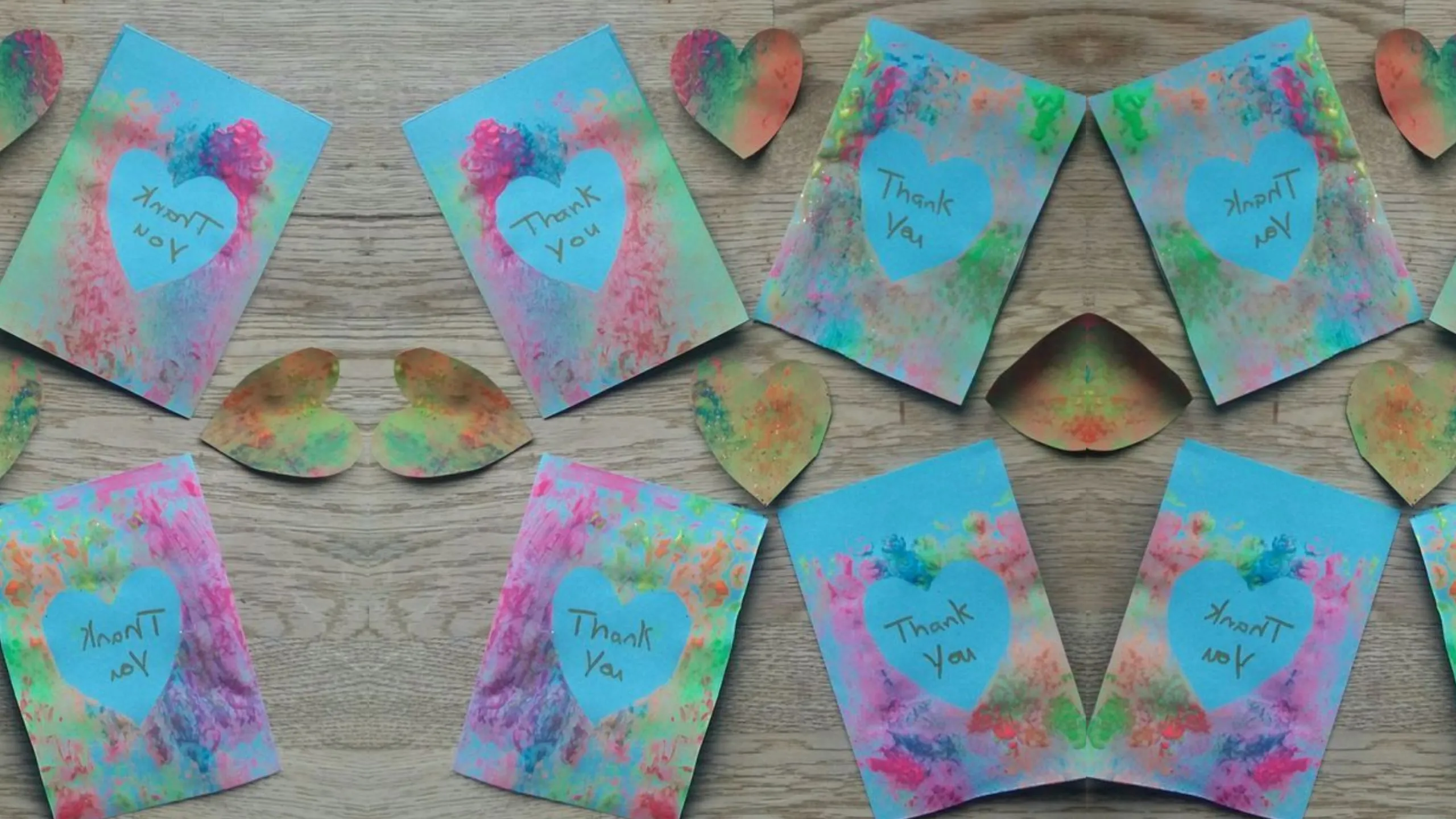 Handmade watercolor thank-you cards with blue heart centers and handwritten messages are displayed on a wooden surface. Bright pink, green, and blue paint accents decorate each card.